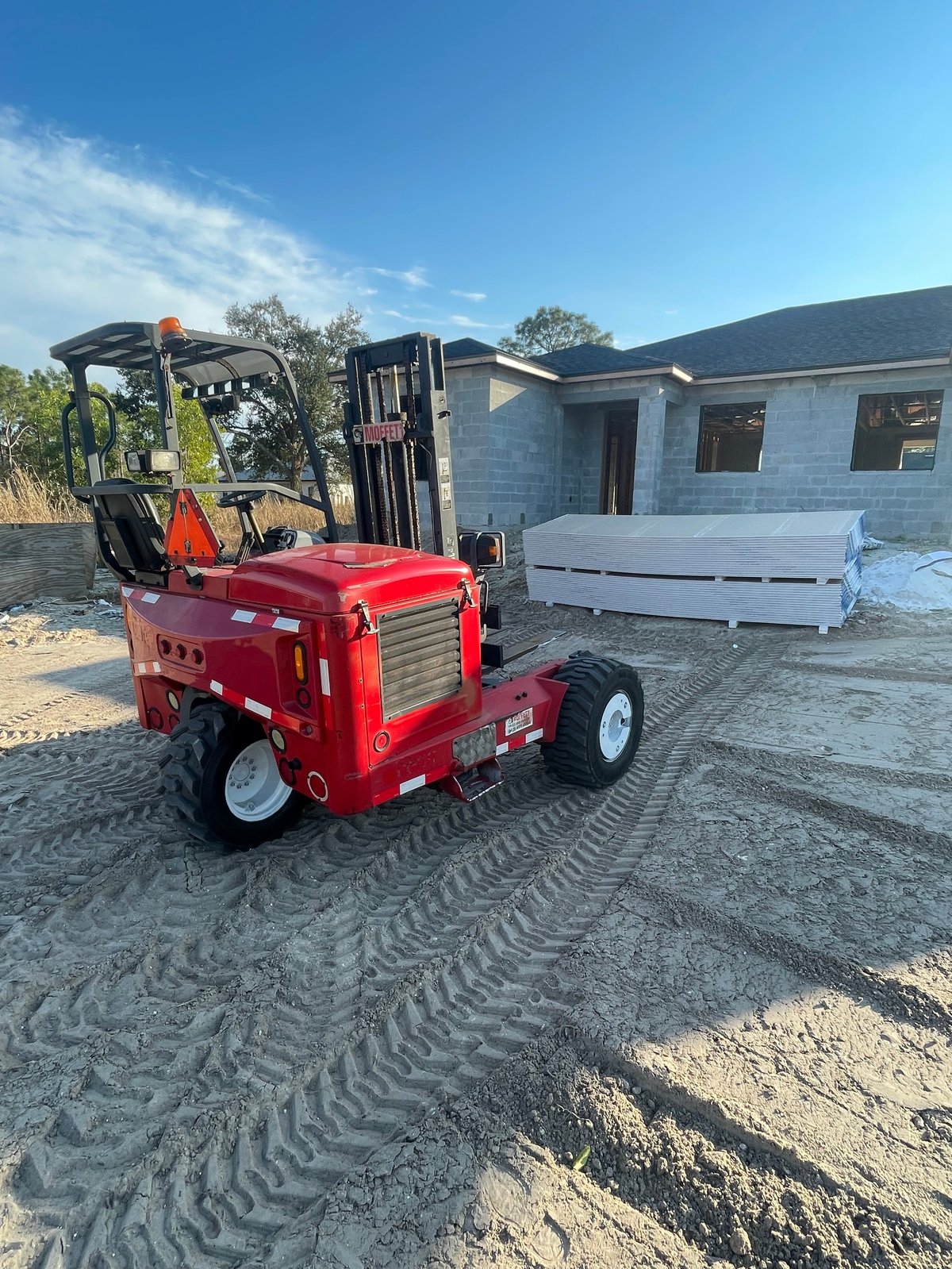 Red compact forklift parked on sandy ground in front of an under-construction house on a clear sunny day