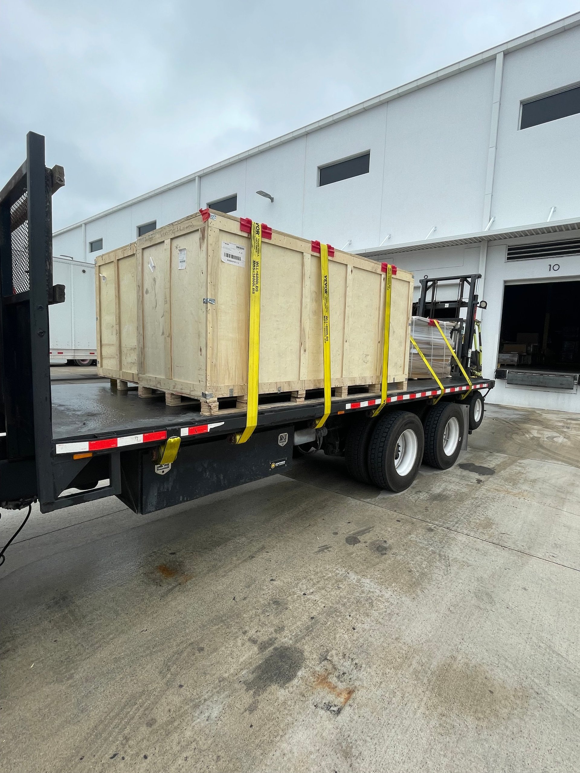 Commercial truck with wooden cargo container secured with yellow straps parked outside industrial warehouse building
