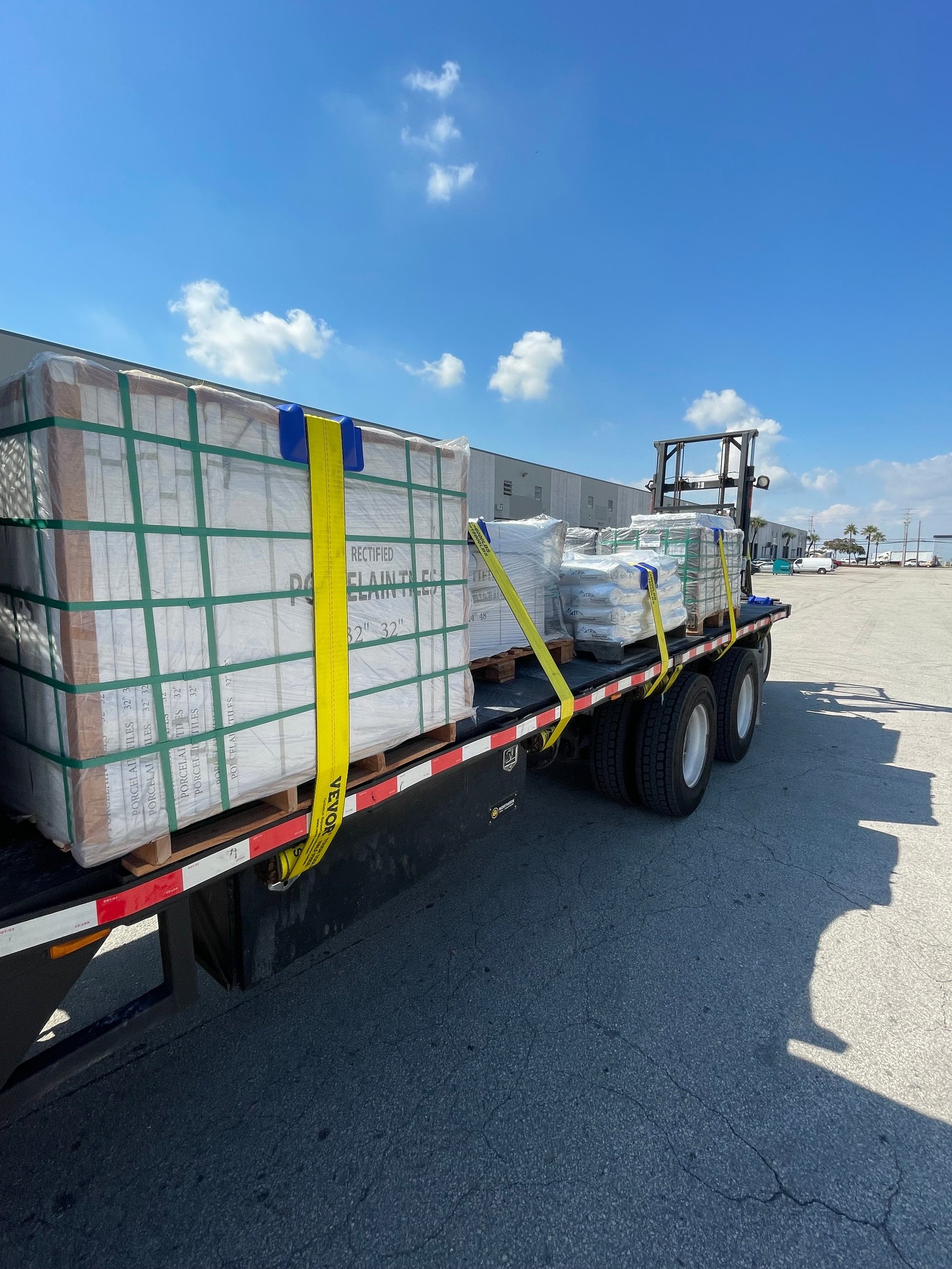 A flatbed truck loaded with large wrapped cargo containers secured with yellow straps in an industrial parking lot