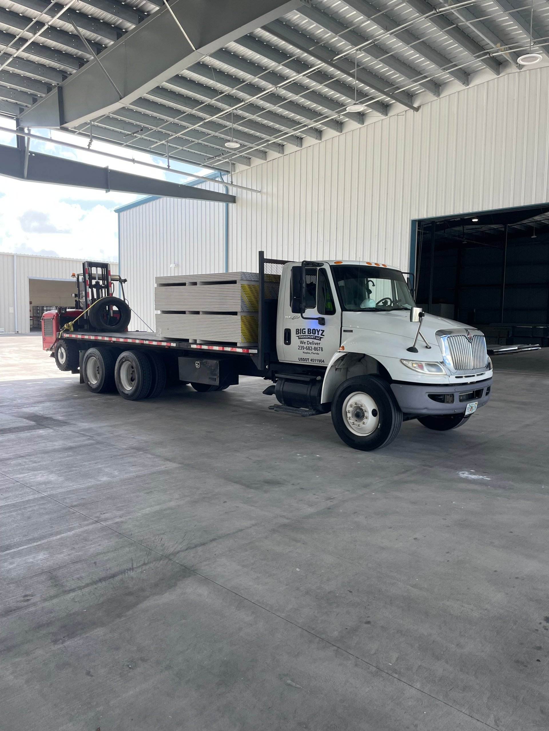 White International truck with flatbed carrying forklift in industrial warehouse with metal ceiling and concrete floor